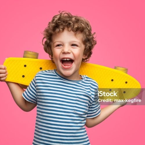 Happy curly boy laughing and holding bright colored skateboard over vibrant pink background isolated